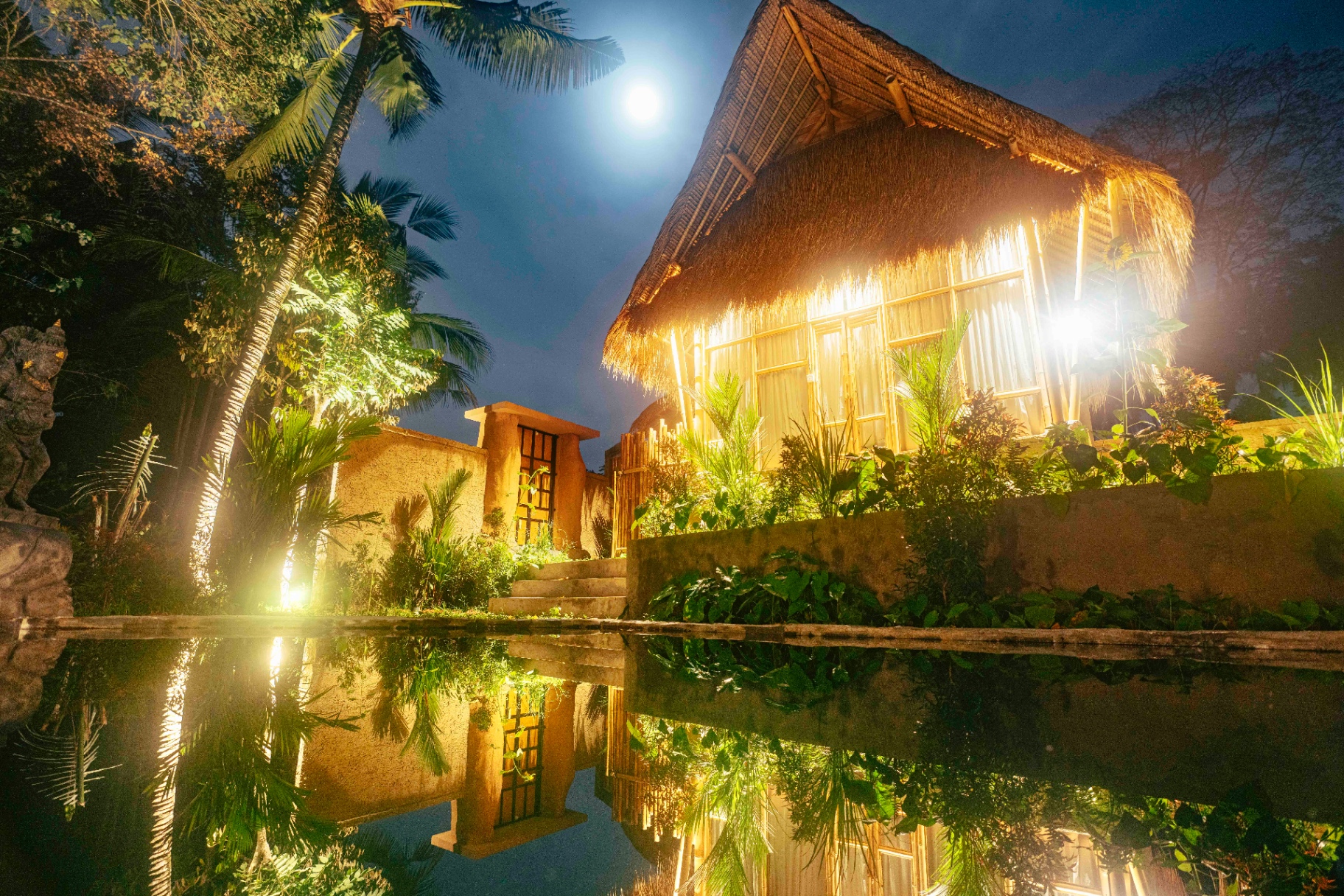 Beleuchtete Villa mit Palmdach bei Nacht, gespiegelt im Pool